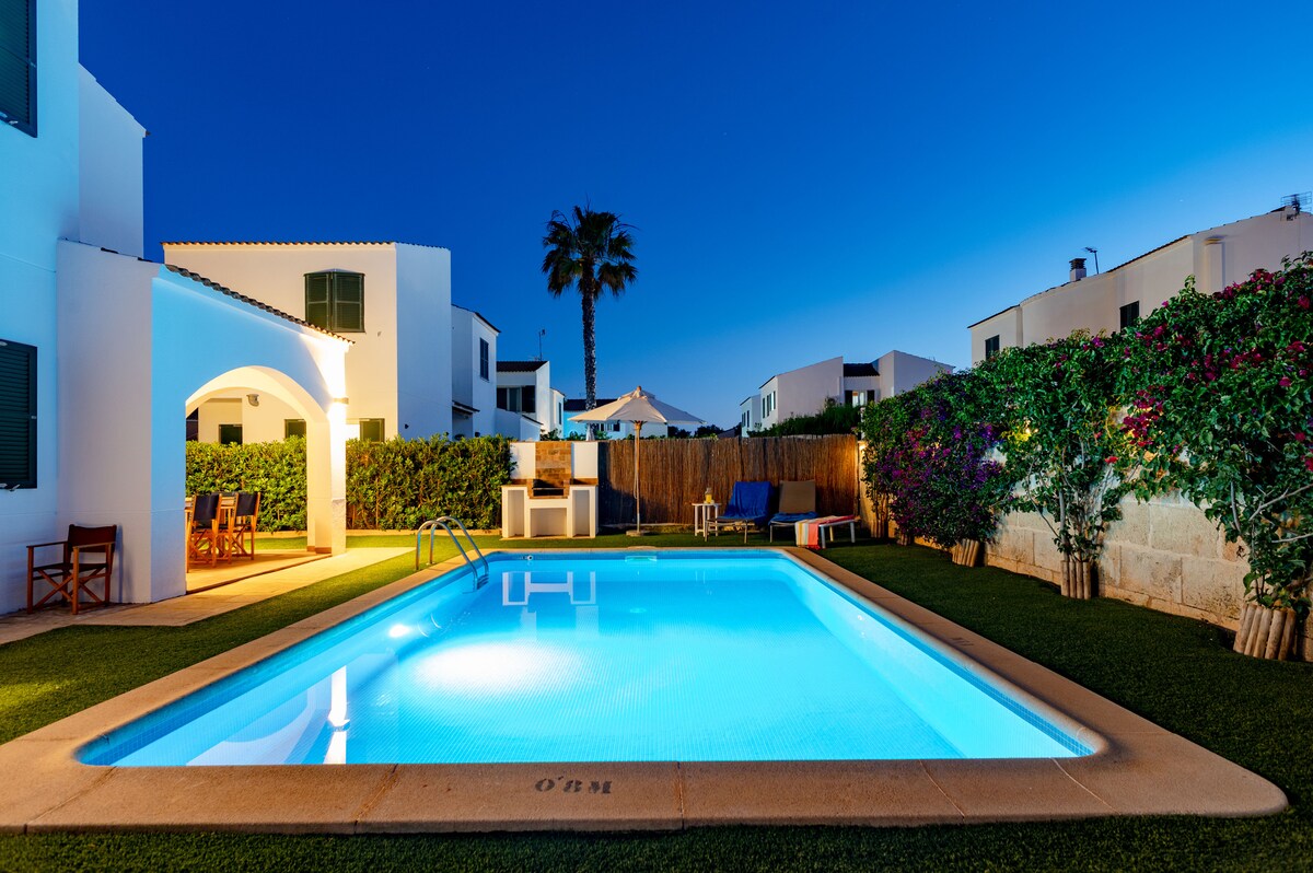 A private pool is surrounded by lush greenery and a well-maintained garden. A barbecue area and outdoor seating can be seen under the soft evening sky. The pool, illuminated in blue, invites relaxation in a peaceful setting.
