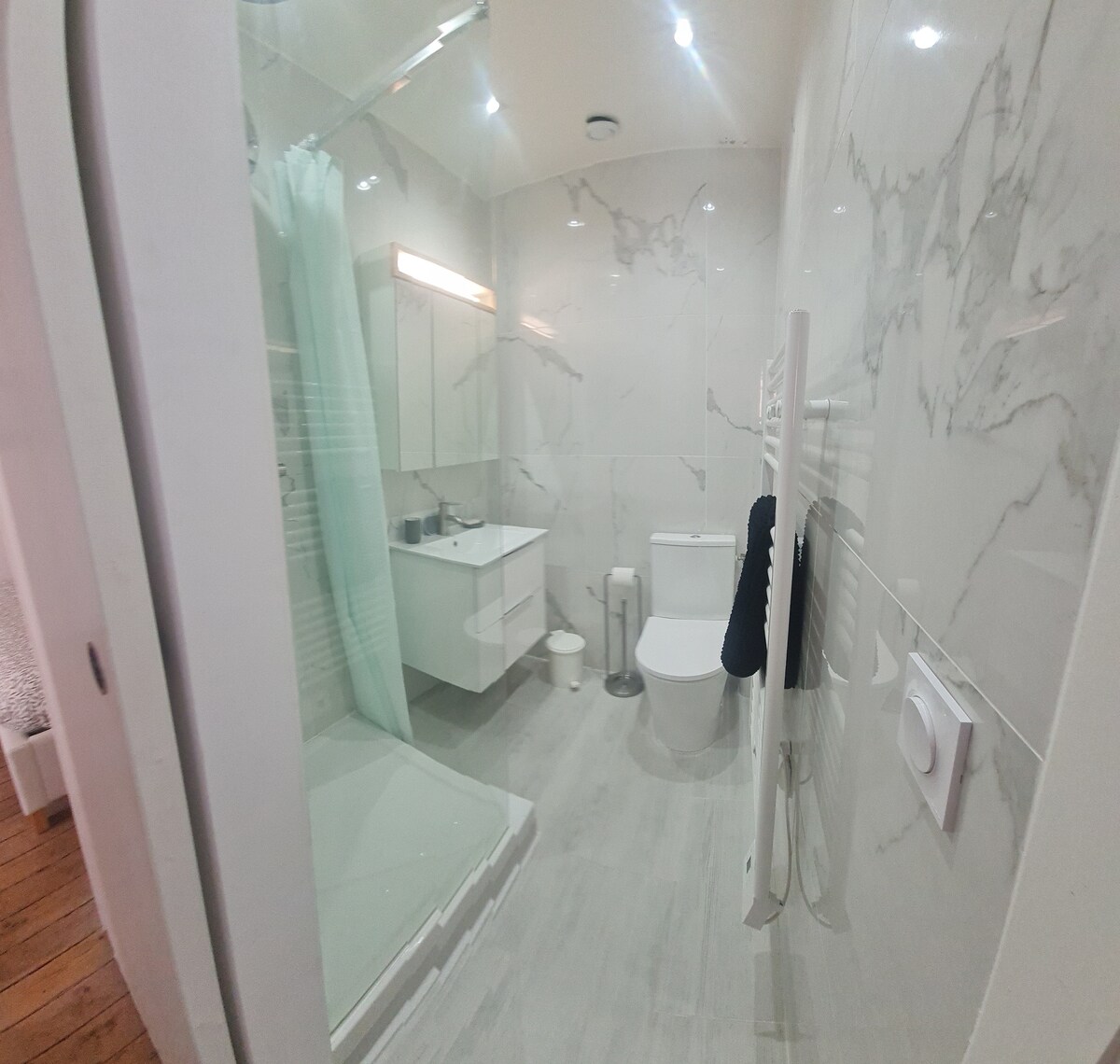 A modern bathroom is presented with marble walls and sleek fixtures. A spacious shower is enclosed with glass, while a white vanity and toilet complete the space. Soft lighting enhances the clean, minimalist design, and towels are neatly hung for convenience.