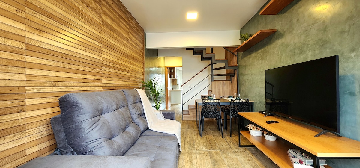 A living area is presented with a comfortable gray sofa, complemented by a wooden accent wall and green-toned walls. A large television is positioned on a wooden media console. A dining table with chairs is visible, along with a staircase leading to the upper level.