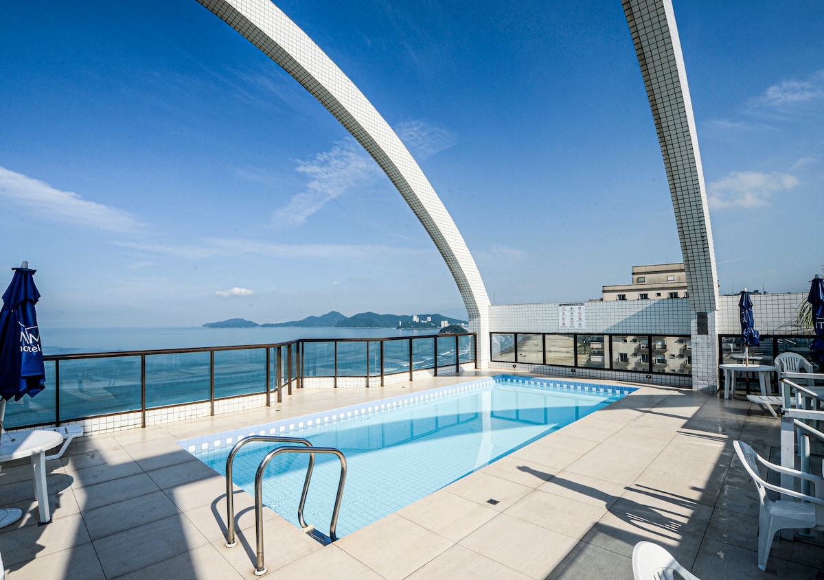 A rooftop pool offers scenic views of the coastline under a clear blue sky. Surrounding lounge chairs and umbrellas provide seating for relaxation. The pool area is designed with modern tiles and features a gentle gradient leading into the water.