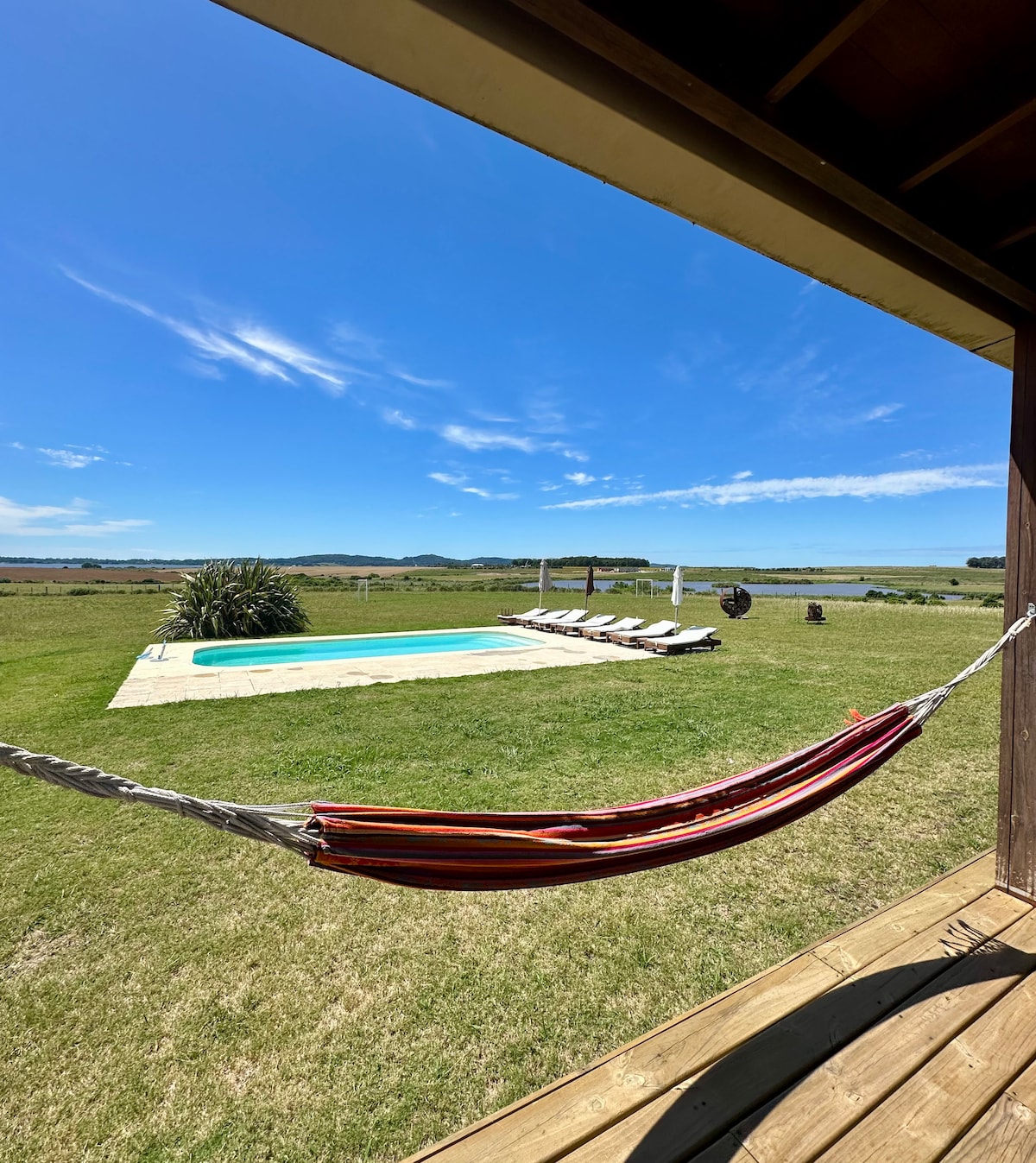 A serene outdoor area is presented, featuring a refreshing swimming pool surrounded by sun loungers on a well-maintained lawn. A colorful hammock hangs nearby, offering a relaxing spot to enjoy the expansive views of the surrounding landscape under a clear blue sky.