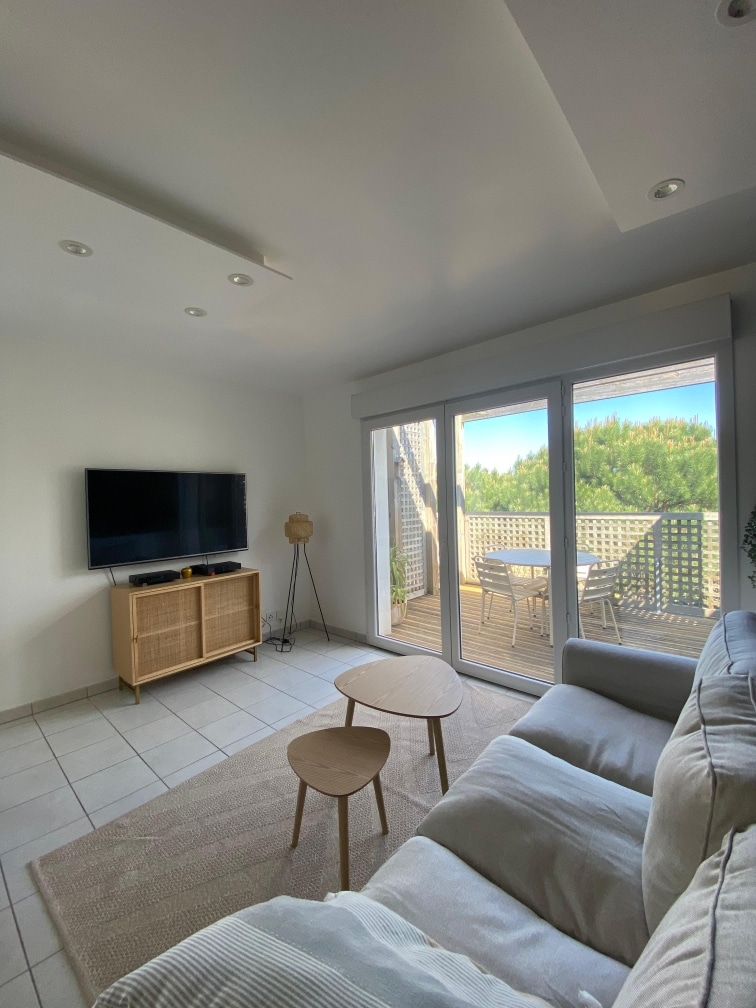 A cozy living area is featured with a light-colored sofa positioned beside two wooden side tables. A television sits on a wooden media unit, while sliding glass doors lead to a sunny terrace overlooking greenery.
