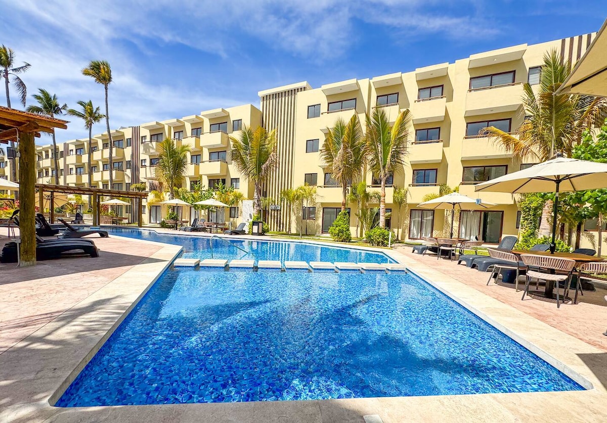 A spacious pool area is framed by palm trees, with clear blue water reflecting the sunny sky. Lounge chairs are arranged around the pool, and several umbrellas provide shade. The modern building is visible in the background, featuring large windows and balconies.