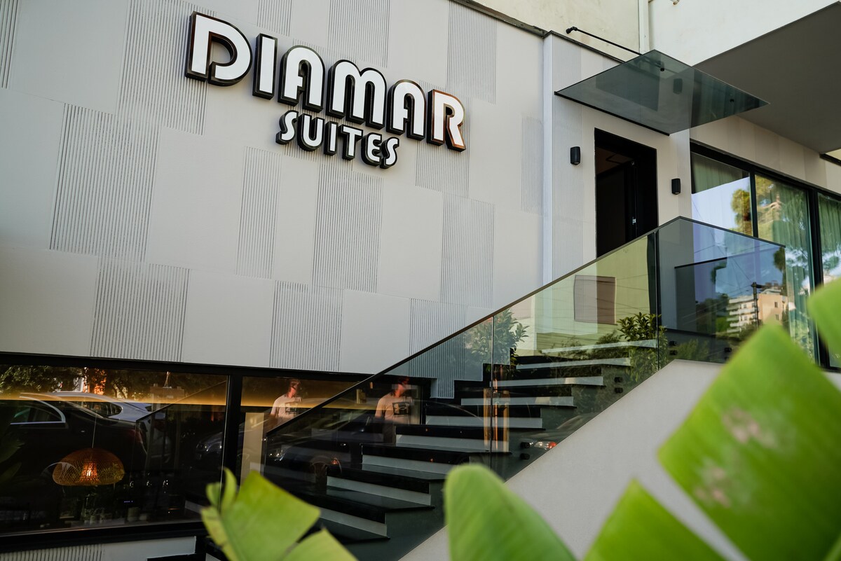 The exterior of the building features the name 'DIAMAR SUITES' prominently displayed above a sleek entrance. A modern staircase with glass railings leads up to the entrance, flanked by lush green plants. Large windows reflect natural light and the surroundings.