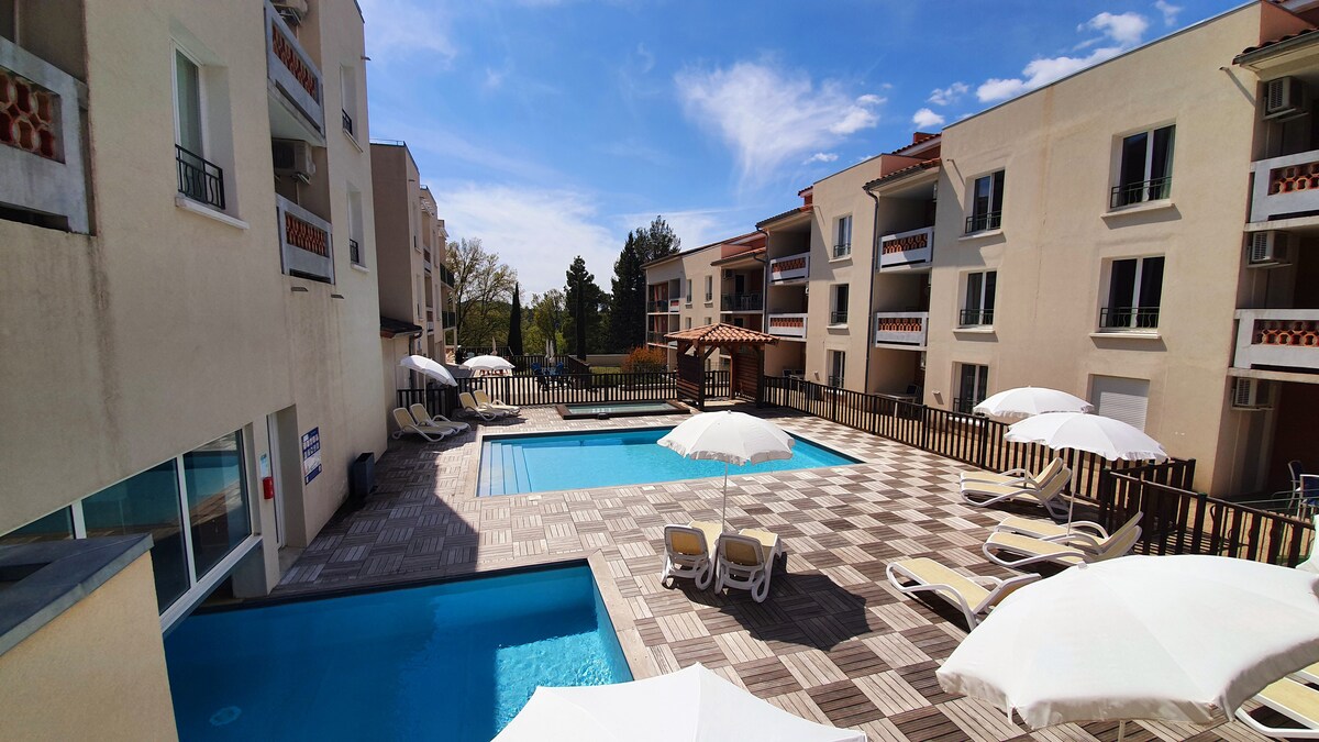A communal outdoor pool area is depicted, featuring two swimming pools bordered by a sun terrace with multiple lounge chairs. Large umbrellas provide shade, and the surrounding modern buildings exhibit a light-colored façade under a clear blue sky.