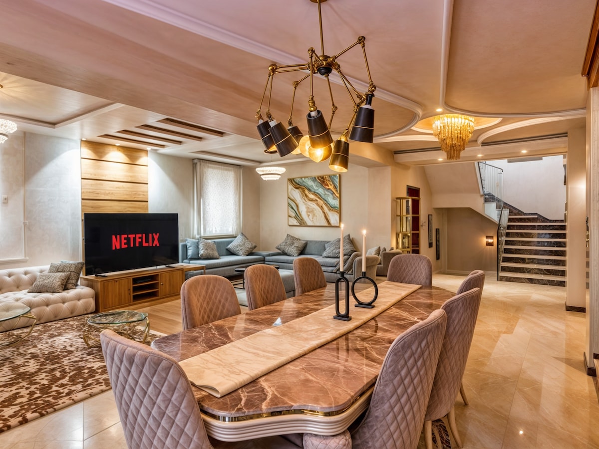 An open-concept living and dining area features a contemporary design with a stylish dining table surrounded by tufted chairs. A large television is mounted on a wall, while a plush sofa complements the space. Elegant lighting fixtures enhance the room's ambiance.