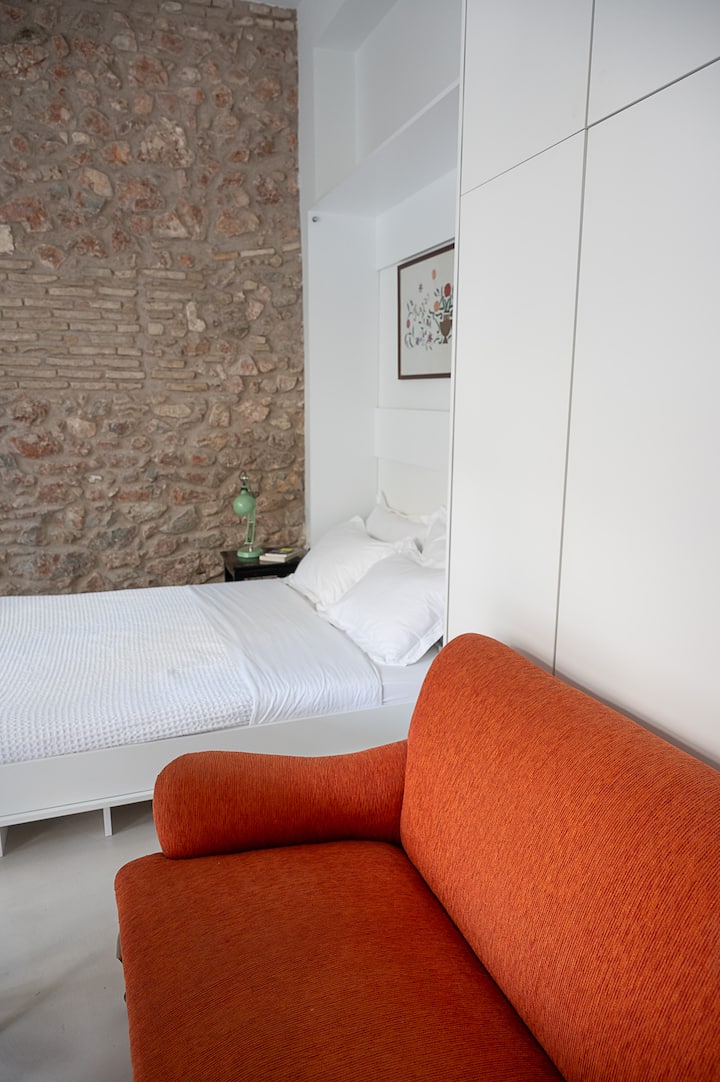 Унифициран простор за спиење и дневен престој во вториот стан, со удобен двоен кревет со бела постелнина и живописен портокалов ретро кауч. Откриениот камен ѕид додава рустикална текстура, додека чистиот бел мебел го балансира просторот со едноставност.