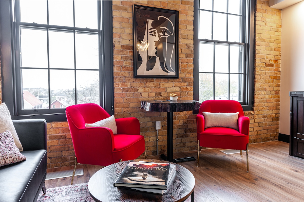 A bright living area features two red chairs positioned around a small round table. Large windows with black frames allow natural light to fill the space. A framed abstract artwork is displayed on the exposed brick wall, adding a touch of modern design.