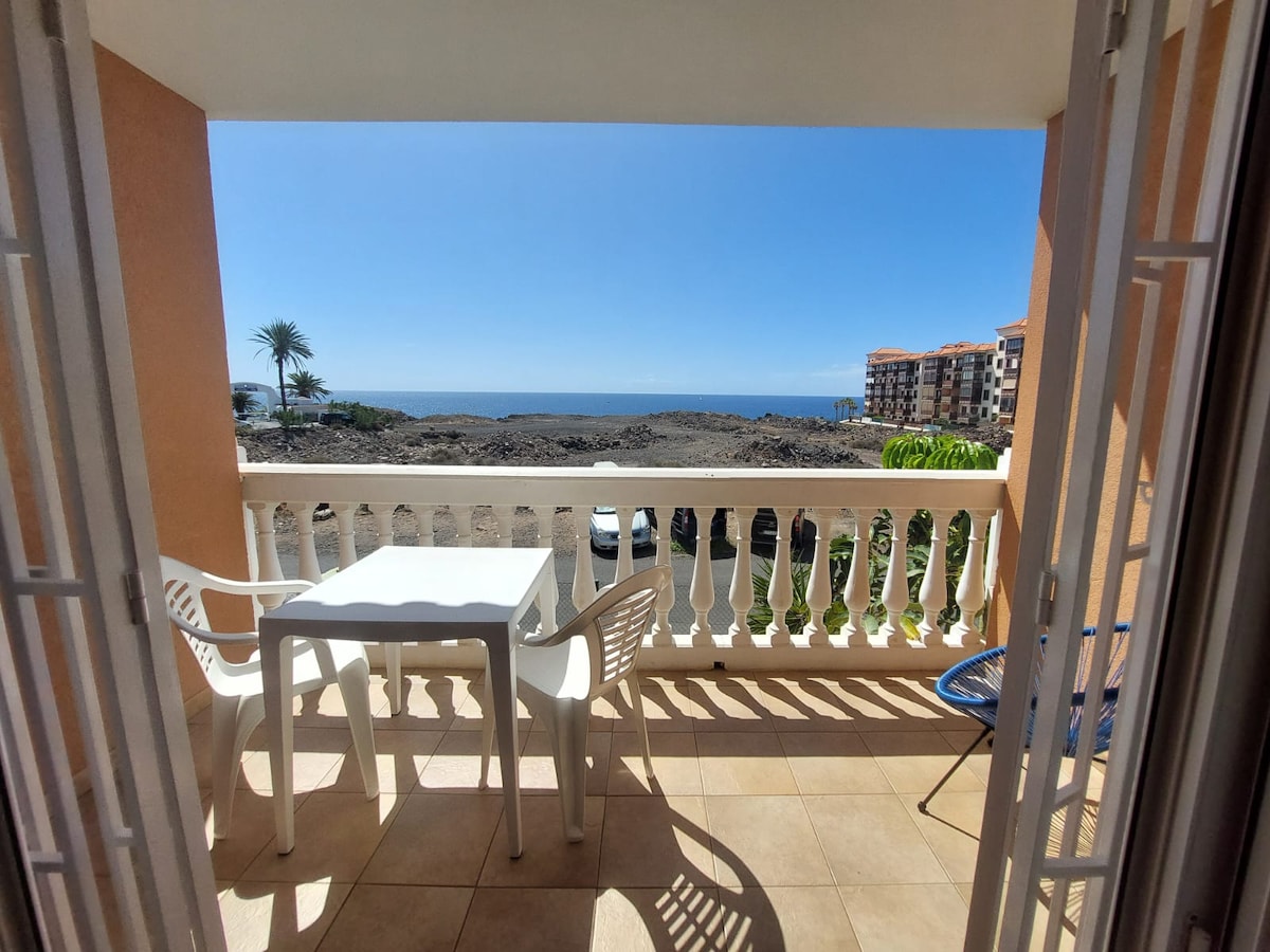 A balcony is shown with a table and chairs facing a scenic view of the sea. Sunlight reflects off the water, and a palm tree is visible nearby. The surroundings include low stone walls and nearby buildings, enhancing the beachside atmosphere.