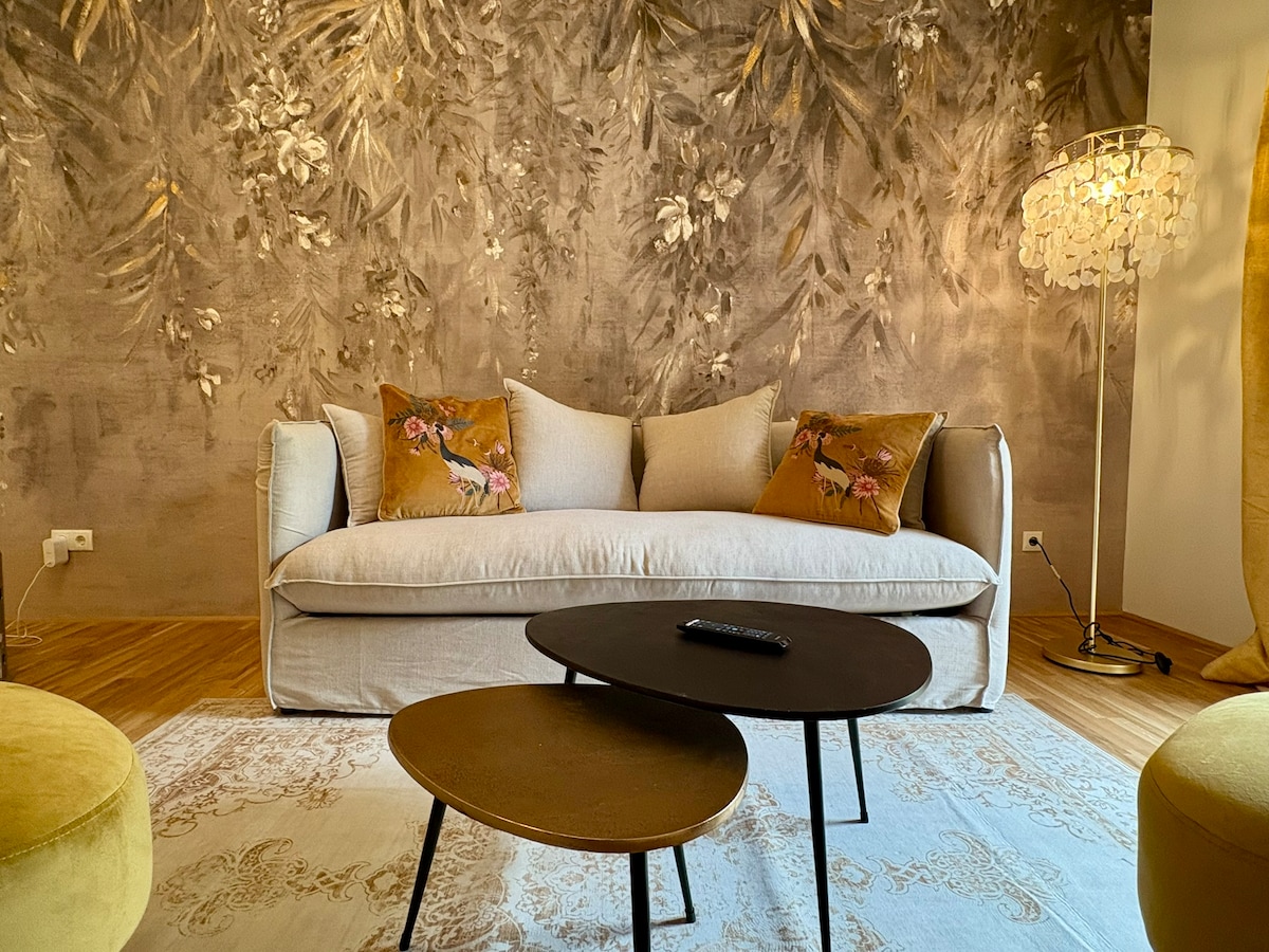A cozy living area is presented, featuring a light-colored sofa adorned with decorative pillows. A pair of small, round tables are positioned in front, with a remote control resting on one. The backdrop showcases a textured floral wall design, complemented by a standing lamp nearby.
