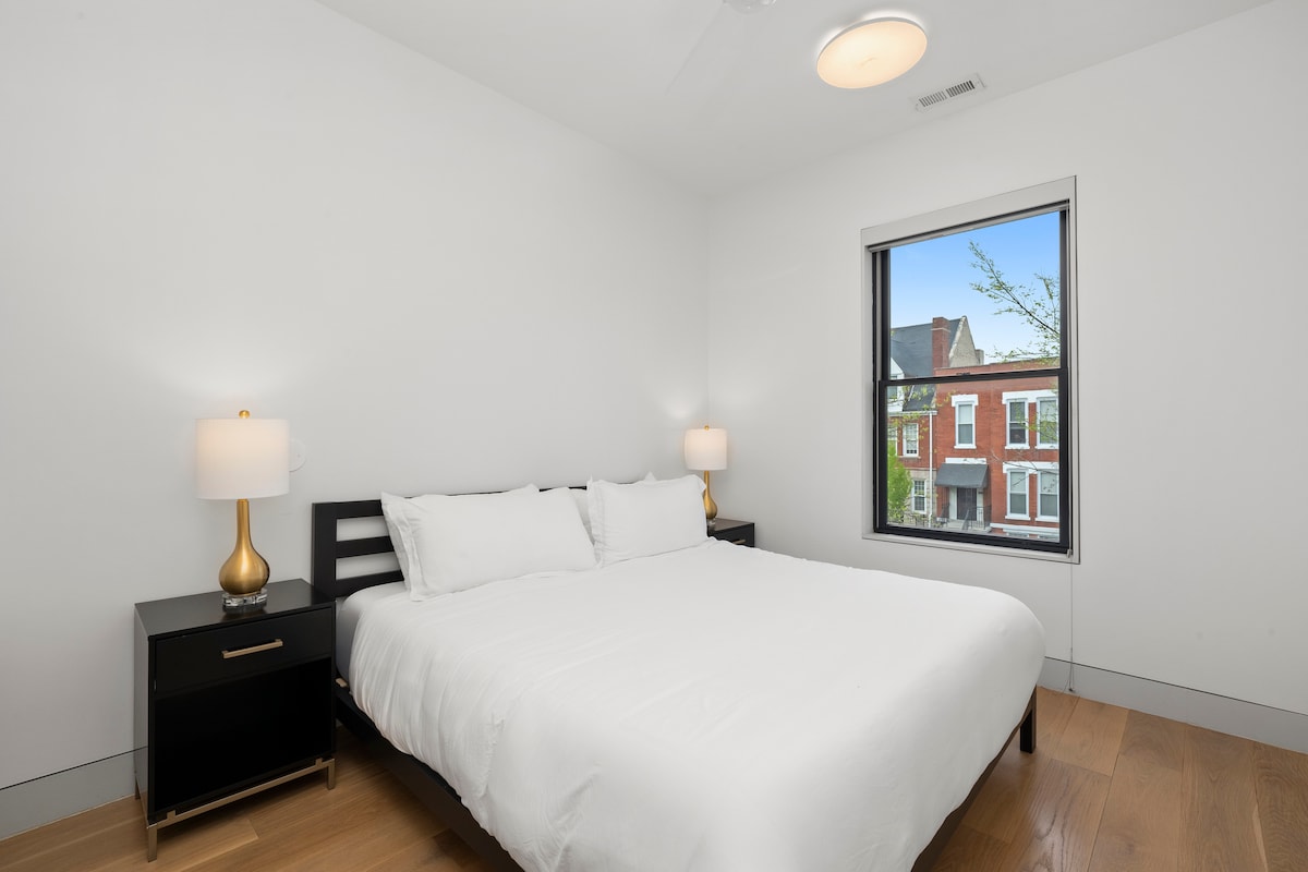 Dormitorio cómodo y elegante con una cama tamaño king, sábanas blancas suaves y mesitas de noche negras elegantes. La gran ventana ofrece hermosas vistas al vecindario y luz natural, perfecta para relajarse y recargar energías durante tu estancia.