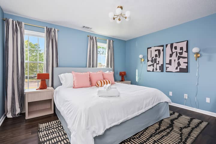 Bright, airy, and full of charm—this king bedroom features statement lighting, bold artwork, and tons of natural light. Blush pillows, striped curtains, 65-inch Smart TV and warm wood tones give it the perfect mix of playful and polished.