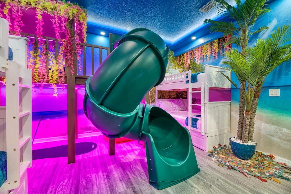 A vibrant gameroom is featured, showcasing a playful green slide extending from a raised platform surrounded by lush decor. Soft lighting in shades of pink and blue creates a cheerful atmosphere, while palm plants add a tropical touch to the inviting space.