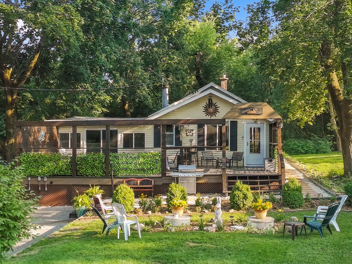 The exterior of a charming house is surrounded by lush greenery, featuring a welcoming porch with seating. A neatly landscaped yard showcases a variety of plants and decorative elements. A path leads toward the house, enhancing the inviting outdoor space.