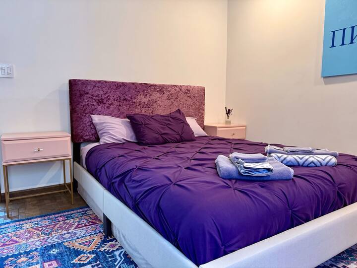 Questa accogliente camera da letto con letto a due piazze presenta una testiera in velluto, biancheria da letto viola e comodini rosa. Un tappeto colorato aggiunge personalità, rendendolo un rifugio elegante e confortevole per notti riposanti o mattine rilassanti.