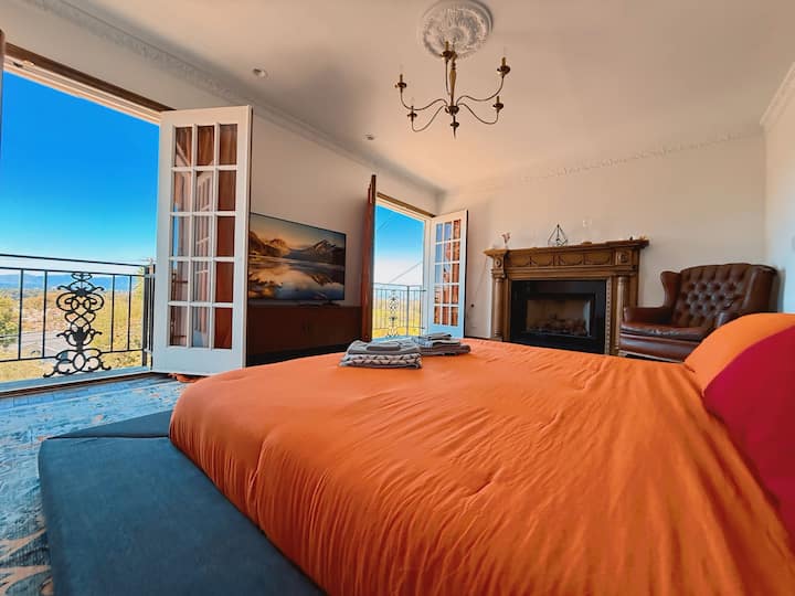 Camera da letto principale con letto king size e portefinestre che si aprono su ampie viste sulle montagne e sulla valle. Dispone di una smart TV da 65", un camino d'epoca e una sedia da lettura in pelle, ideale per serate accoglienti o per svegliarsi con la luce dorata del sole californiano.