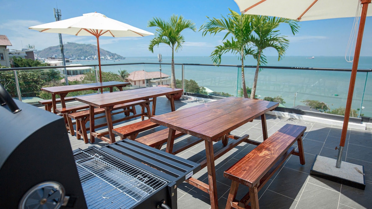 A large rooftop terrace is featured, equipped with wooden picnic tables and benches under white umbrellas. A barbecue grill is positioned nearby. A panoramic view of the ocean and distant hills is visible, offering an inviting outdoor space suitable for gatherings.