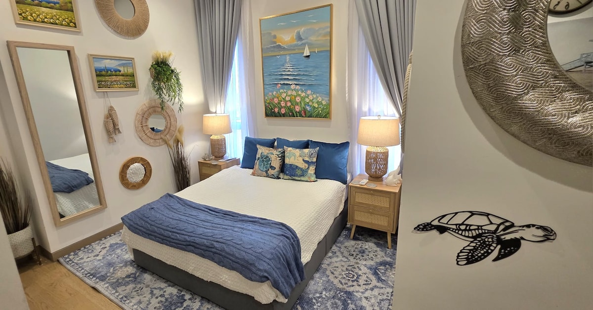 The bedroom features a spacious queen bed adorned with decorative pillows and a blue blanket. Soft lighting is provided by bedside lamps. Framed artwork of a serene waterscape adds a calming touch. Natural elements are incorporated with greenery and decorative mirrors on the walls.