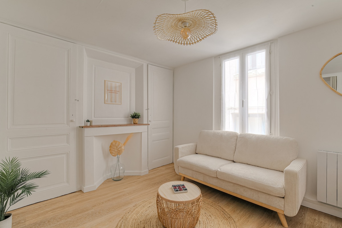 A bright living area features a light, neutral-toned sofa positioned next to a round coffee table on a wooden floor. A decorative wall mirror and a small plant add subtle touches to the minimalist design, complemented by natural light entering through two elegant windows.