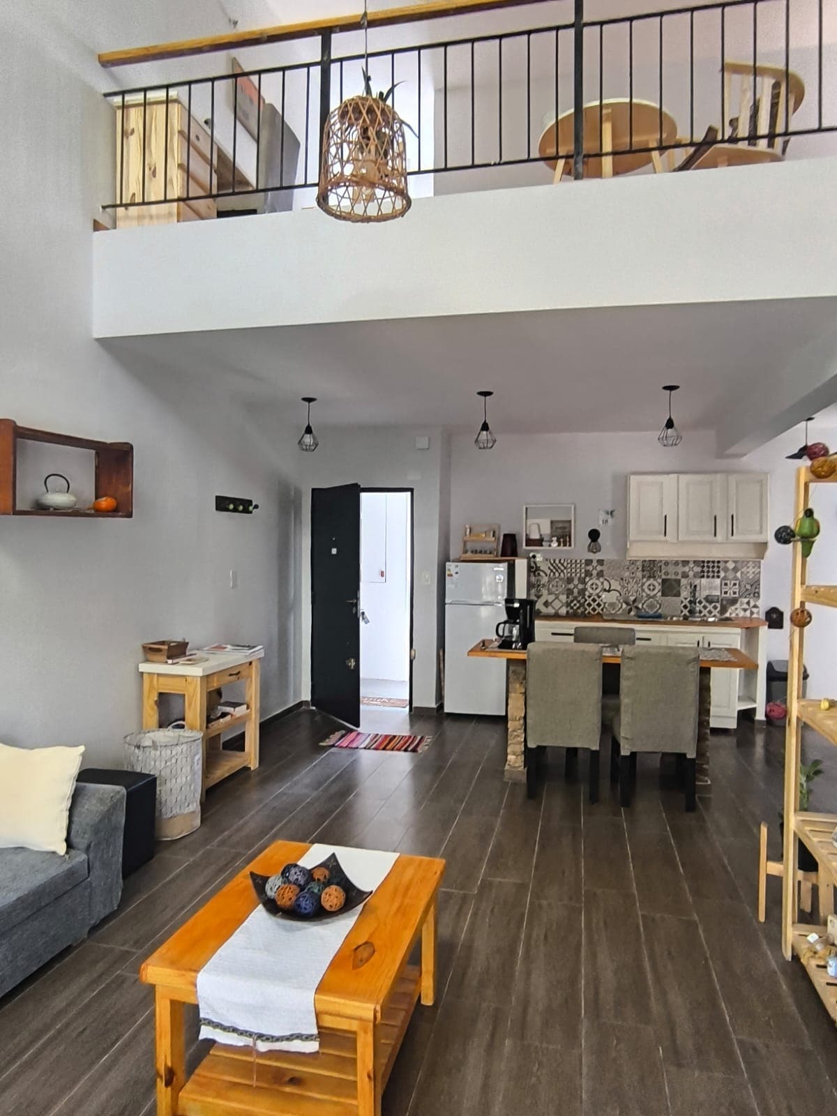 An open living area features a dark grey sofa and a wooden coffee table with decorative items. A dining space with grey upholstered chairs can be seen beside a kitchen with light-colored cabinetry and patterned backsplash. The ceiling is accented by pendant lights and a charming hanging lamp.