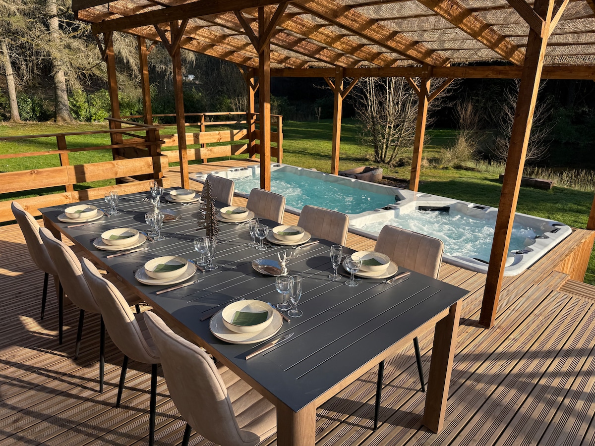 An outdoor dining area features a large wooden table set with elegant dishware and glassware. A spacious hot tub and a swim spa are situated nearby, surrounded by lush greenery. The shaded pergola provides a comfortable space for gatherings in a serene natural setting.