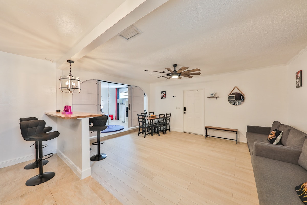 An open living area is presented, featuring a light-colored wood floor and a modern design. A small dining table sits beside a gray sofa, and black bar stools are arranged near a kitchen counter. Bright lighting enhances the spacious atmosphere.
