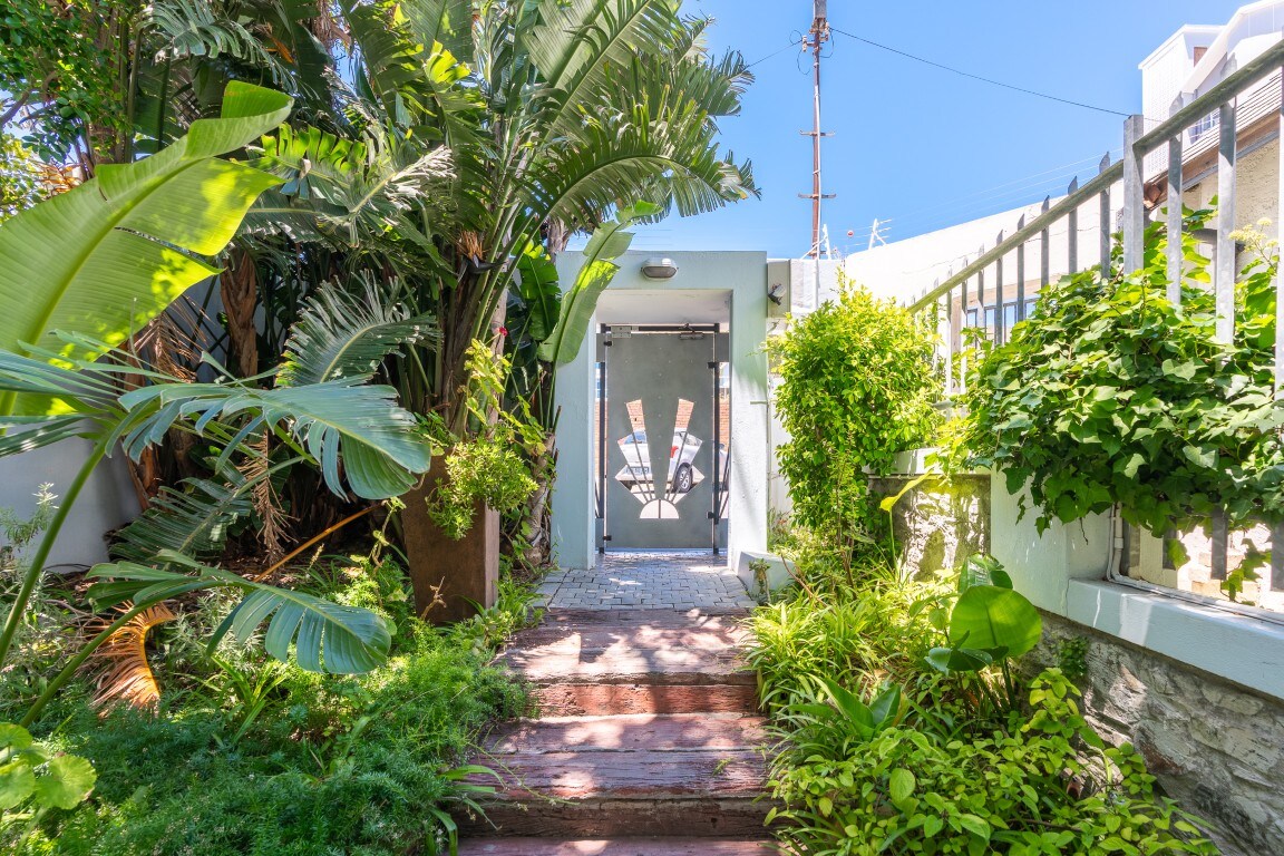 A pathway leads through lush greenery towards an entrance adorned with artistic designs. Tropical plants frame the entrance, contributing to a serene and welcoming ambiance. The sunlight filters through, highlighting the natural beauty of the surroundings.