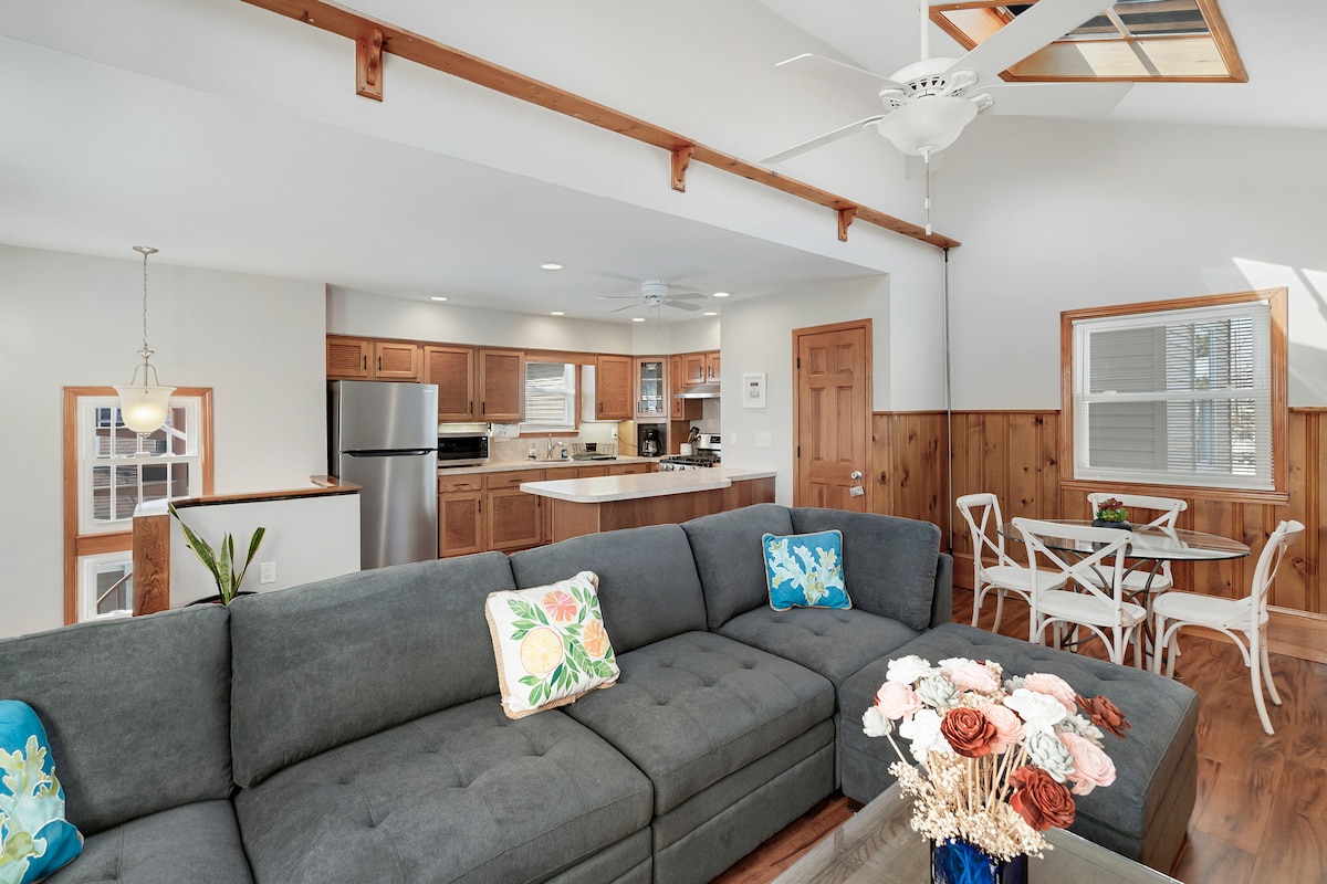 A spacious living area features a large gray sectional sofa adorned with colorful throw pillows. Natural light fills the room, highlighting the open layout that includes a modern kitchen with stainless steel appliances and a dining area with a small white table and chairs.