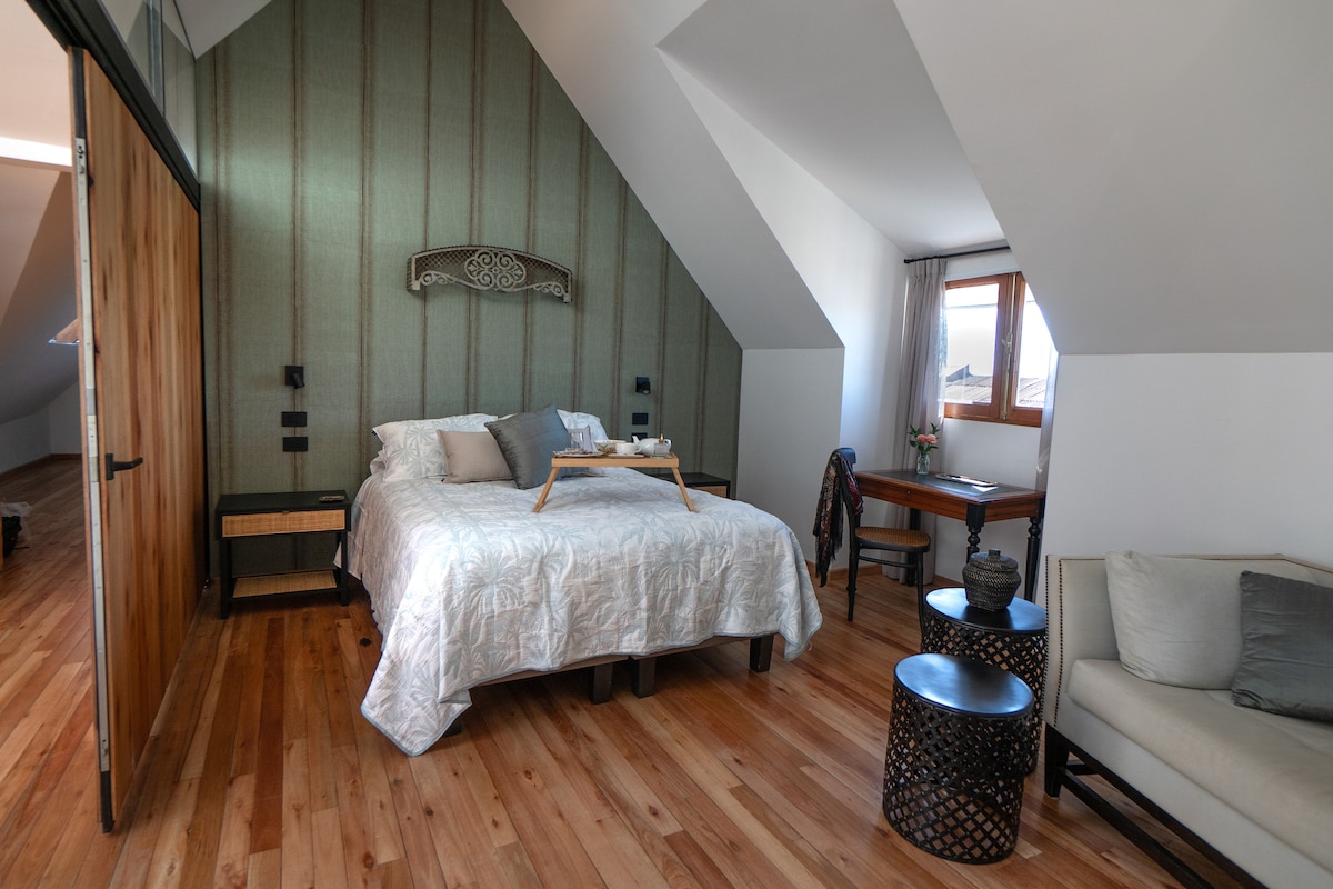 An airy bedroom features a spacious bed with a textured white coverlet, complemented by decorative pillows. A small breakfast tray rests atop the bed. Nearby, a wooden desk and chair are positioned against a sloped wall, with natural light filtering through a window.