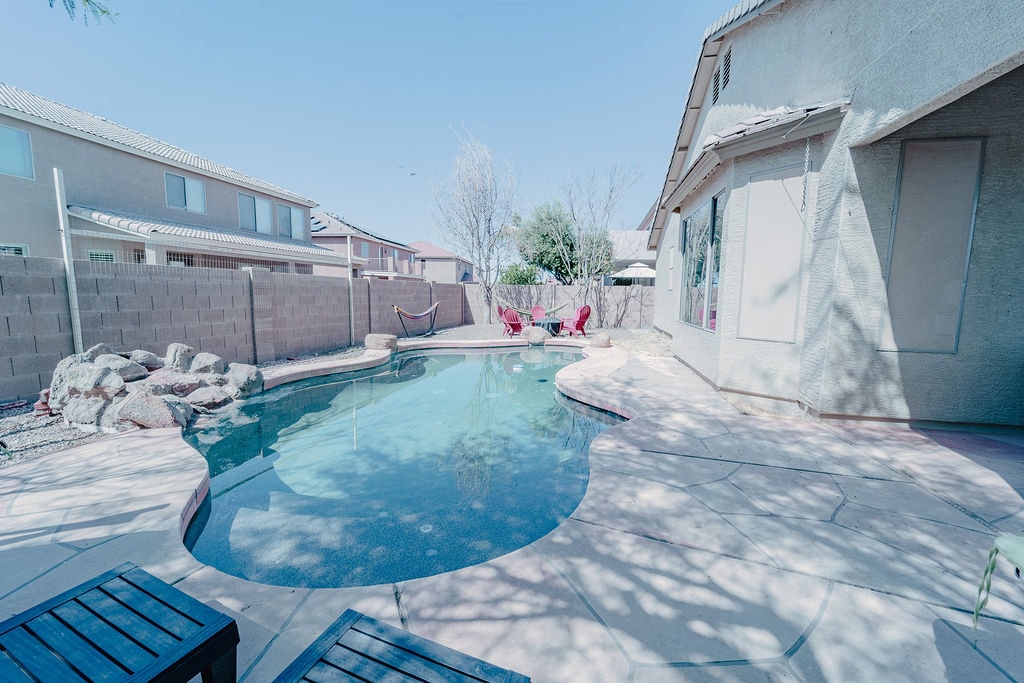 A serene outdoor space features a sparkling pool surrounded by a smooth stone patio. Two red chairs are placed nearby, with a hammock visible in the background. Lush greenery provides a natural touch, enhancing the tranquil atmosphere.