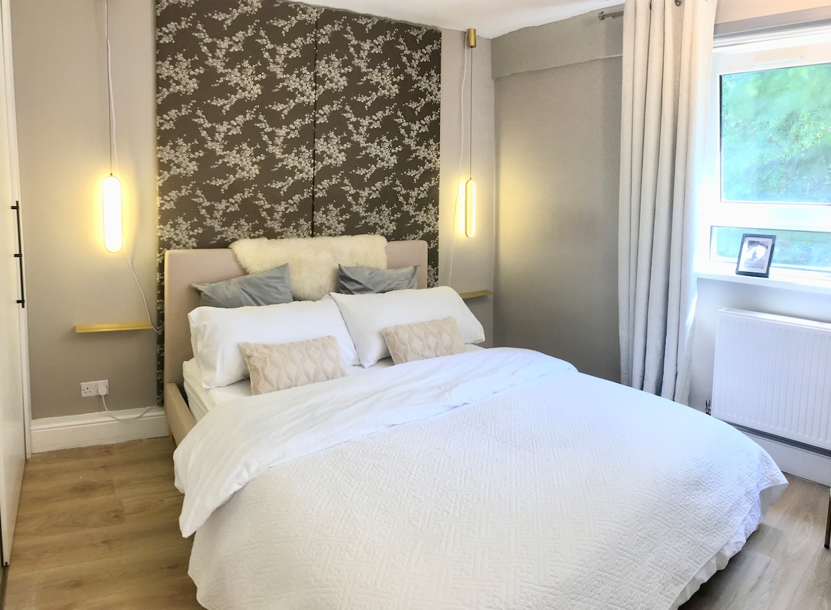 A well-appointed bedroom features a spacious bed dressed in white linens, complemented by two pillows and decorative cushions. Soft lighting is provided by wall-mounted fixtures, and a window allows natural light to illuminate the space, with a view of greenery visible outside.
