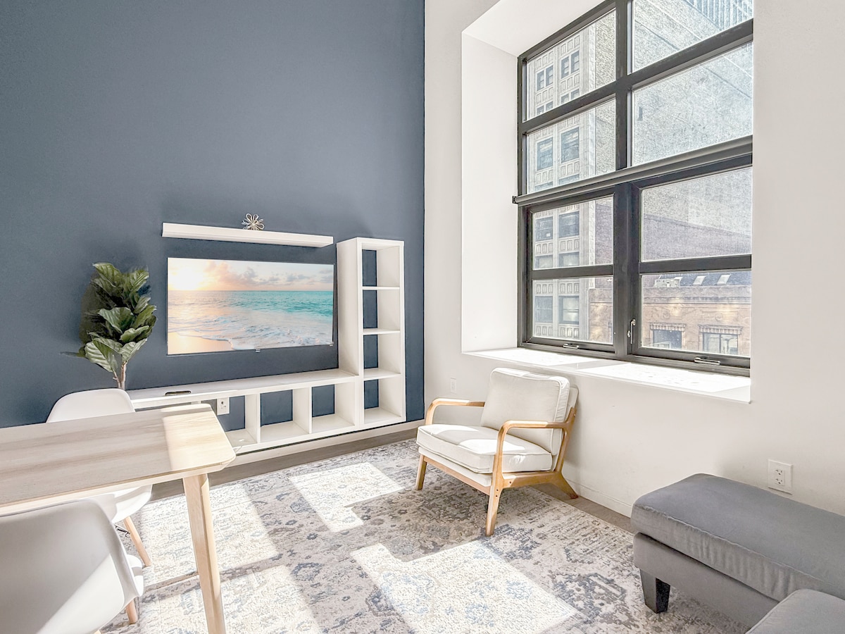 A bright living area features large windows allowing natural light to fill the space. A comfortable chair and an ottoman are arranged next to a dining table. A wall-mounted TV displays a serene beach scene, complementing the neutral color palette of the room.