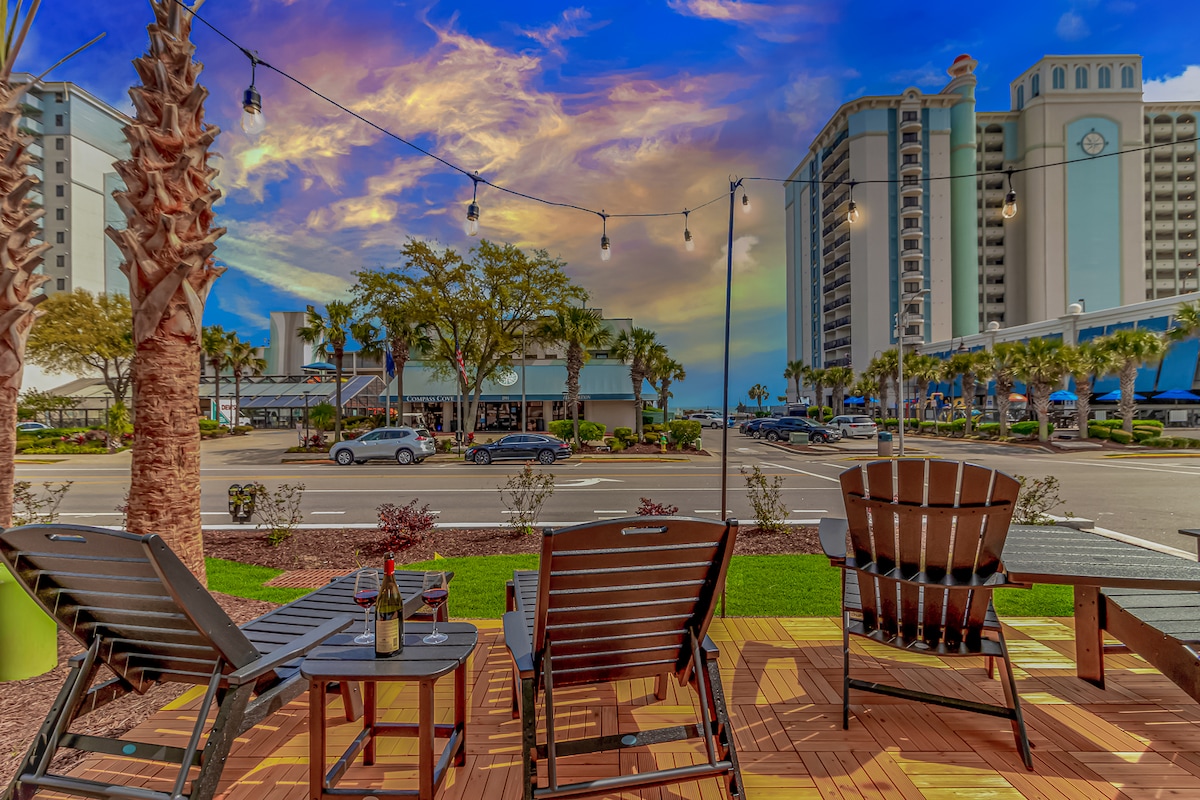 A welcoming outdoor patio area is presented with brown lounge chairs set against a backdrop of palm trees and colorful sky. Strings of lights are visible above, and the nearby buildings highlight a vibrant coastal atmosphere. The space encourages relaxation and enjoyment of the surroundings.