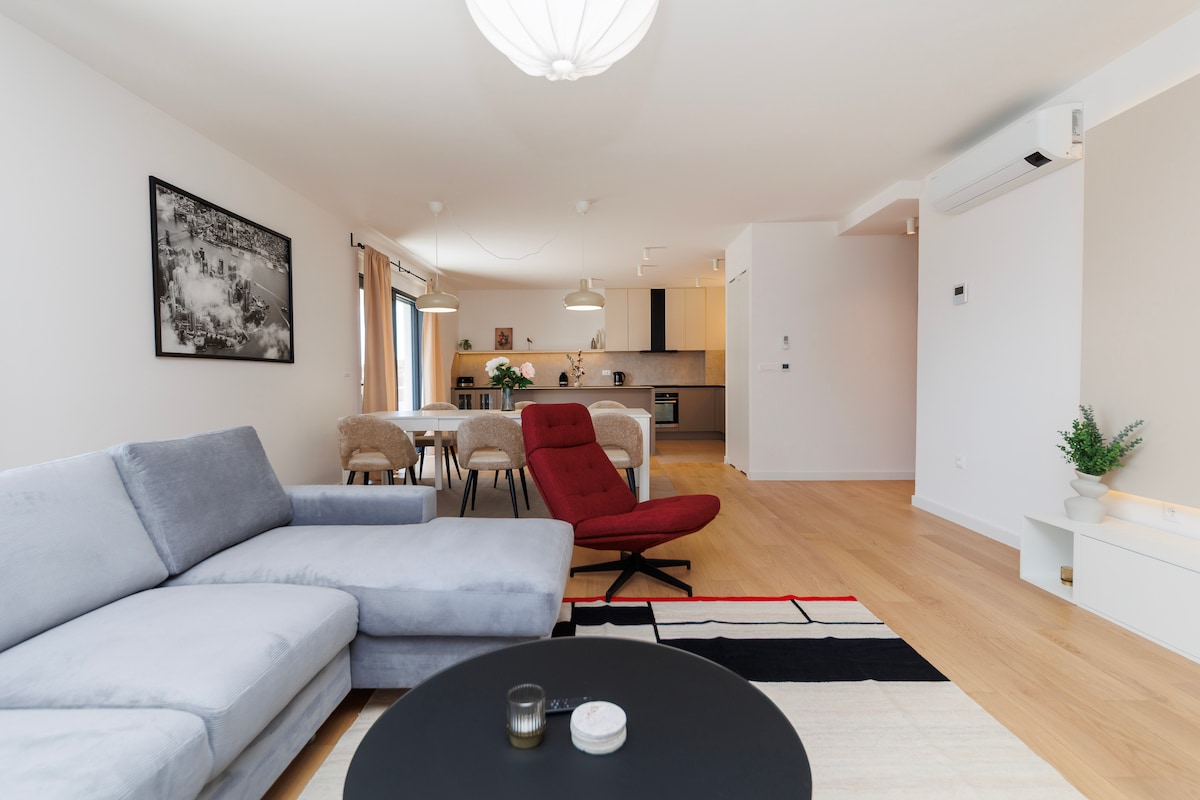 The spacious living area features a light grey sectional sofa paired with a bold red chair, set against a soft area rug with geometric patterns. A modern coffee table holds a candle, while a dining area with plush chairs and elegant lighting is visible in the background.