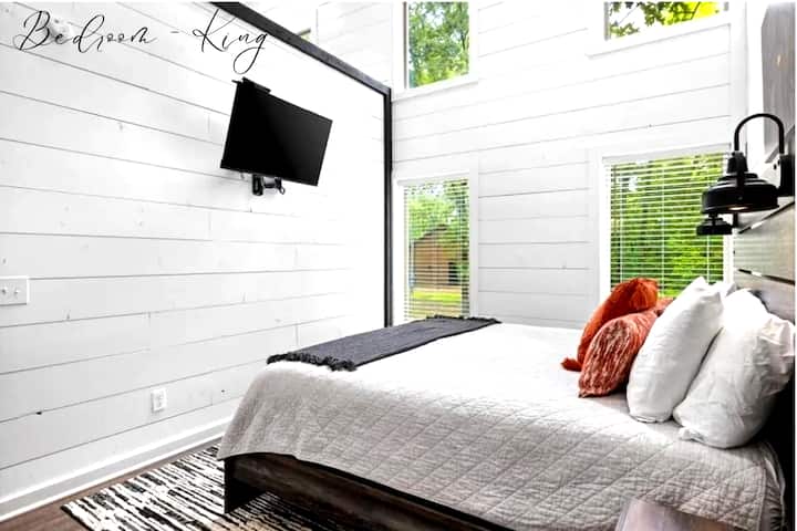 Светла спалня за гости с телевизор с плосък екран и много прозорци.