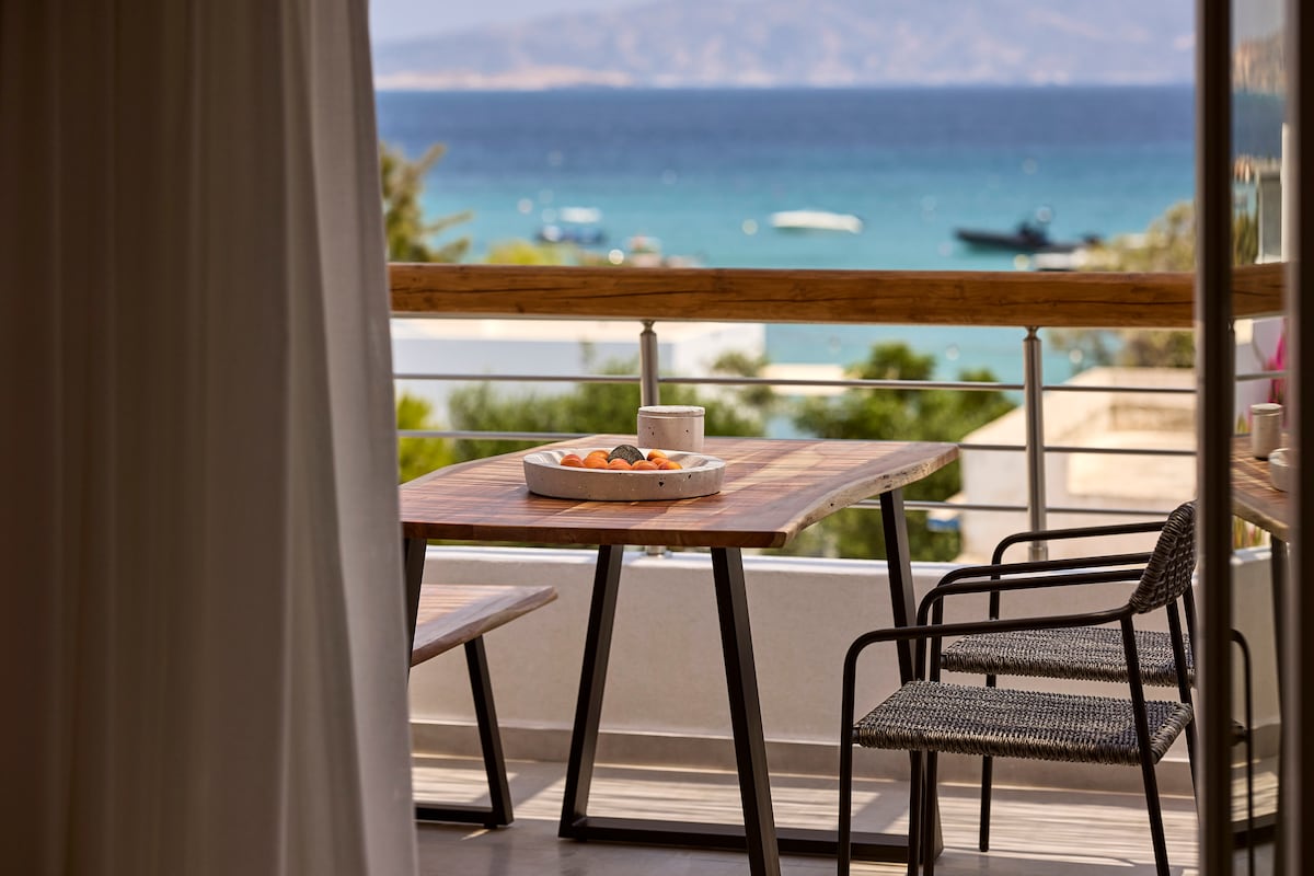 A wooden dining table is positioned on a balcony, accompanied by two dark chairs. A bowl of fruit sits at the center of the table. The background features a serene view of the sea and distant hills under a clear sky.