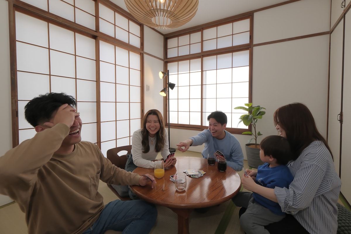 A traditional Japanese-style room features sliding shoji doors and a round wooden table at its center. Four individuals engage in conversation, with drinks placed on the table. A potted plant adds a touch of greenery, enhancing the space’s warm ambiance.