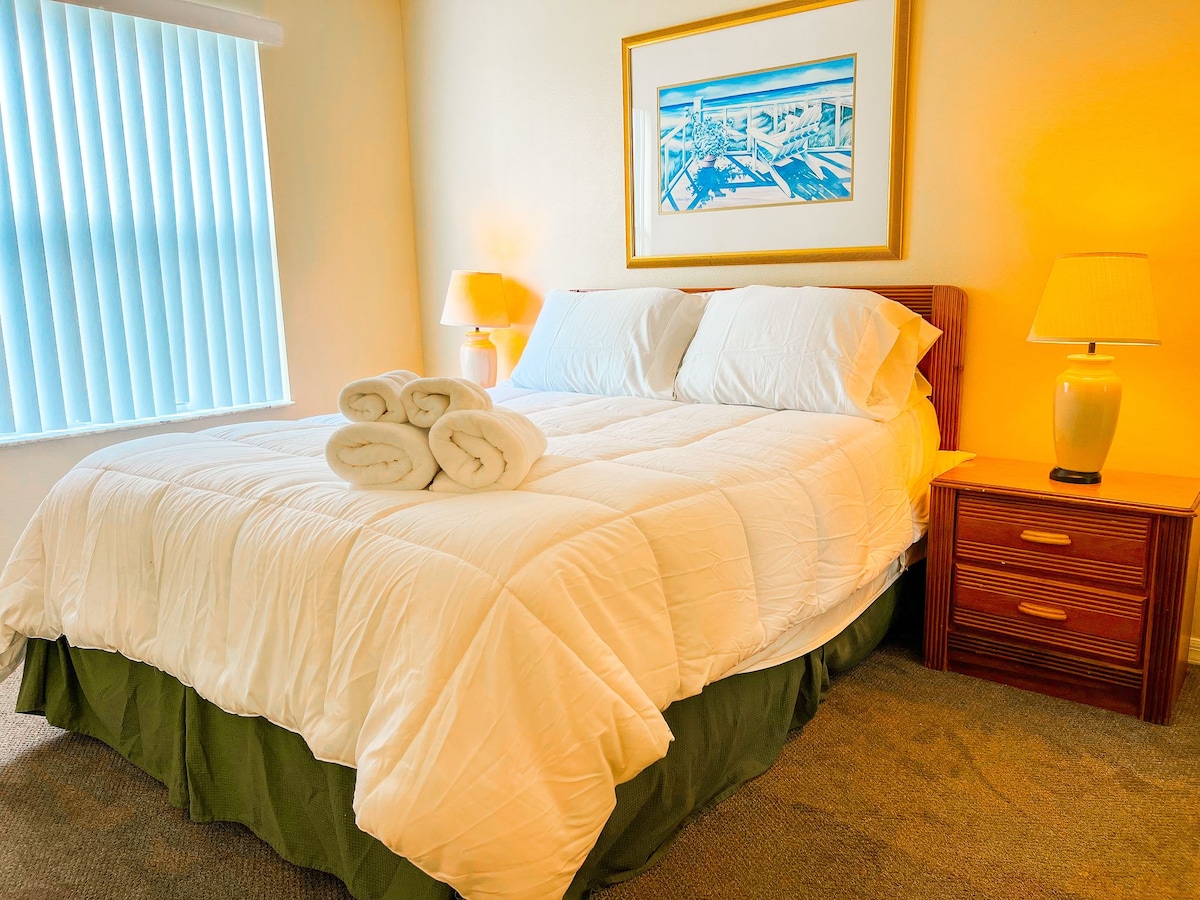 A comfortable bedroom features a spacious bed with white linens and neatly arranged towels. Soft lighting from bedside lamps creates a serene environment. A framed beach scene adorns the wall, while vertical blinds filter natural light from a window nearby.