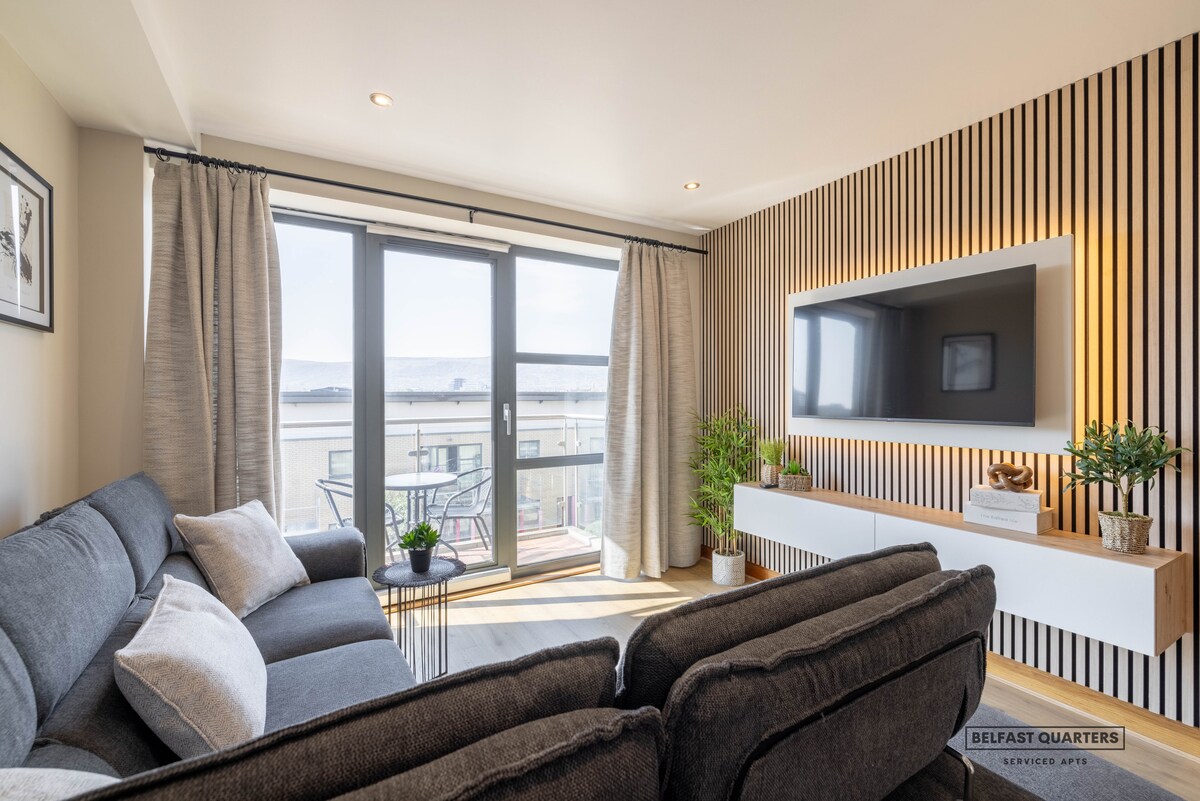 A modern living area features a spacious grey sofa situated across from a wall-mounted TV. Natural light is welcomed through large glass doors leading to a private balcony, while decorative plants add a touch of greenery beside the furniture. Wooden panel accents line the wall.