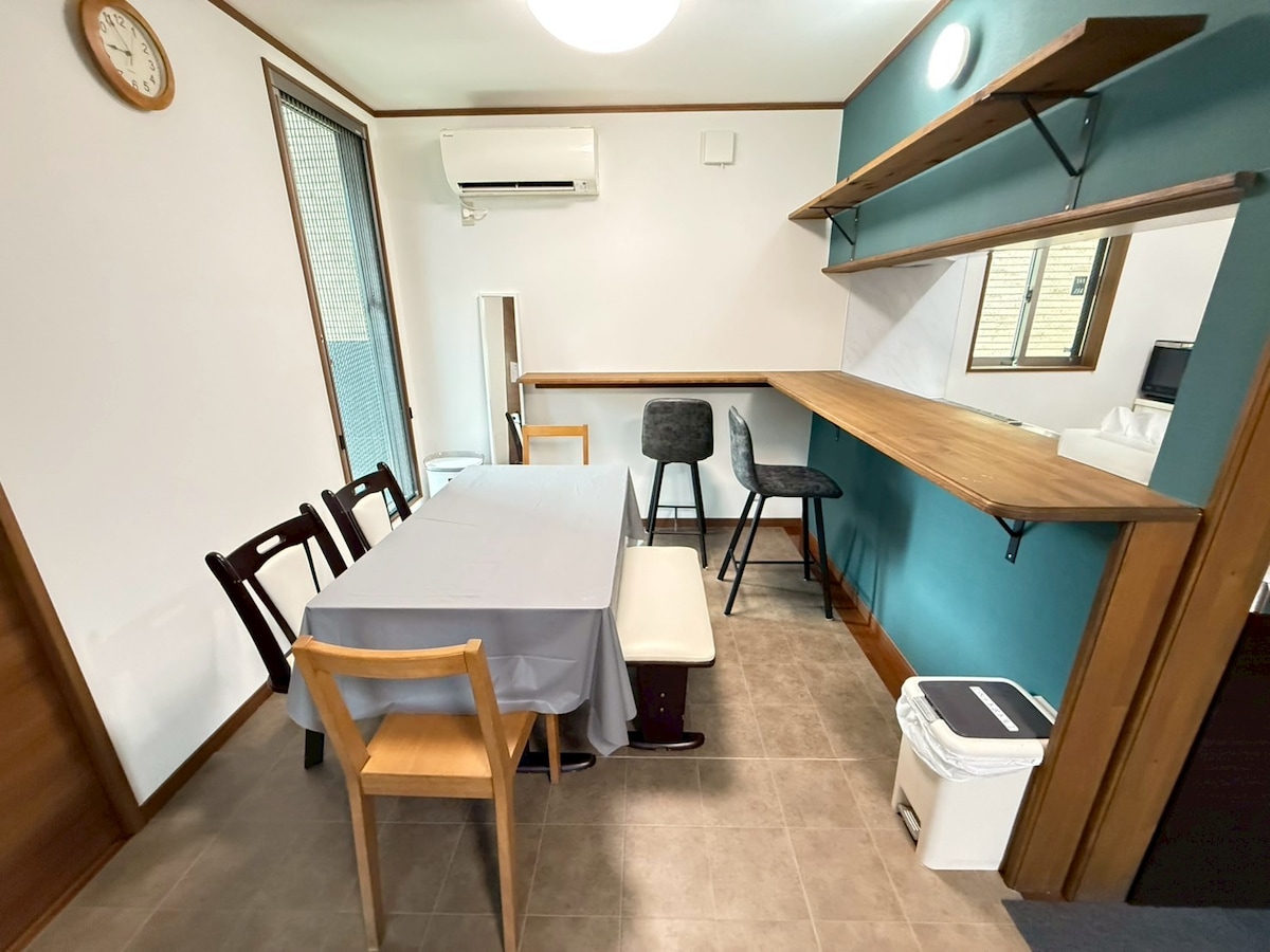 A dining area is presented with a rectangular table covered by a gray tablecloth, surrounded by varying chairs. A compact kitchen counter with two high stools is positioned against a teal wall, accompanied by a mirror and stylish wall shelf above. Natural light is diffused through a window.