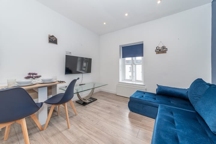 Un salón moderno y vibrante con un lujoso sofá azul, elegantes obras de arte en las paredes y luz natural, perfecto para desconectar después de un día fuera y una televisión para ver películas y relajarse después del trabajo.