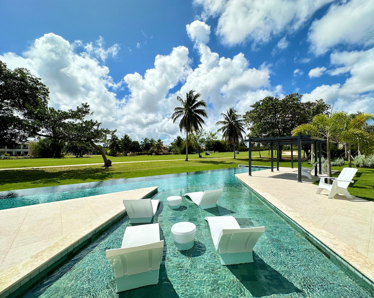 A serene pool area is framed by lush greenery and palm trees under a bright blue sky. Comfortable lounge chairs with accent tables are positioned on a sun shelf, creating a relaxing space for guests. The pool's clear water reflects the surrounding landscape.