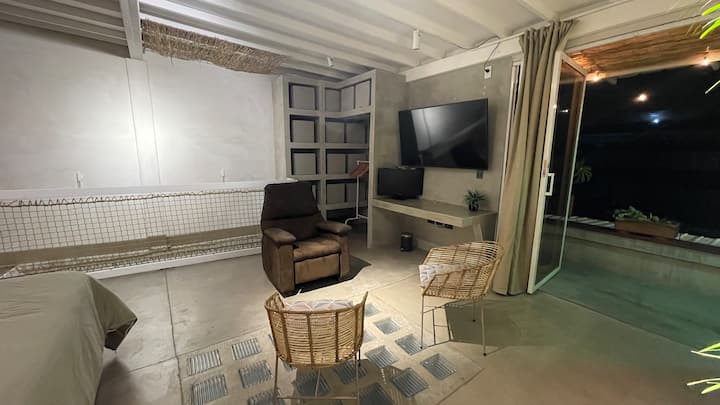 Sala acogedora equipada con televisor, sillón reclinable y sillas de mimbre, ideal para descansar, leer o disfrutar de una película. La conexión con el balcón permite integrar el interior con el exterior, creando un ambiente fresco y relajante.
