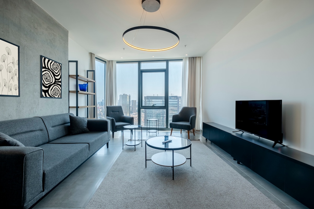 A modern lounge area features a grey sofa and two armchairs, complemented by a round coffee table. Large windows provide an expansive view of the cityscape. A sleek media console holds a television, while a minimalist wall art piece adds a touch of creativity.