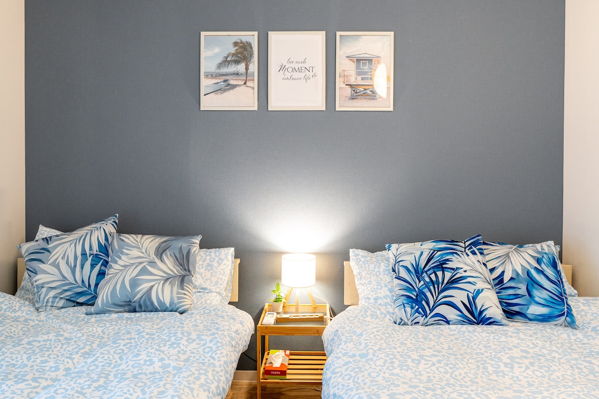 A tranquil sleeping area features two spacious beds with blue and white patterned bedding. Decorative pillows in tropical designs are arranged on the beds. A small bedside table with a lamp is placed between them, complemented by framed art pieces on the wall.