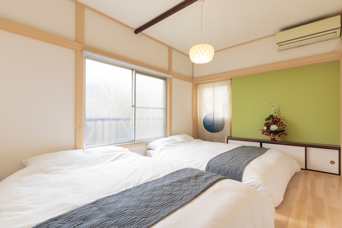A well-lit bedroom features two spacious beds dressed in white linens, with a decorative navy blue runner on each. Natural light enters through a window, and a calming green wall is accented by a simple artwork and a small floral arrangement.