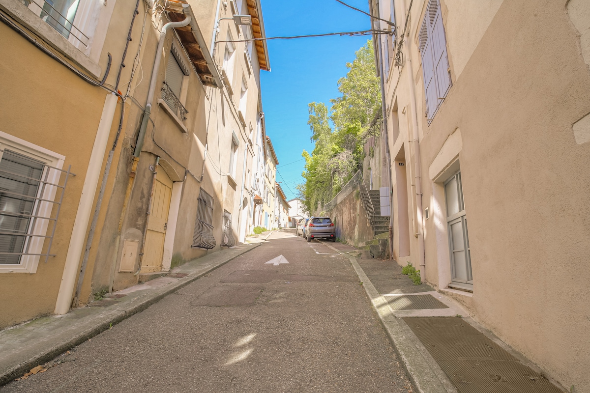 A narrow, quiet street is depicted, lined with light-colored buildings on either side. The road slopes gently upward, leading towards a blue sky. A parked car is visible near the side of the street, contributing to the neighborhood's residential feel.