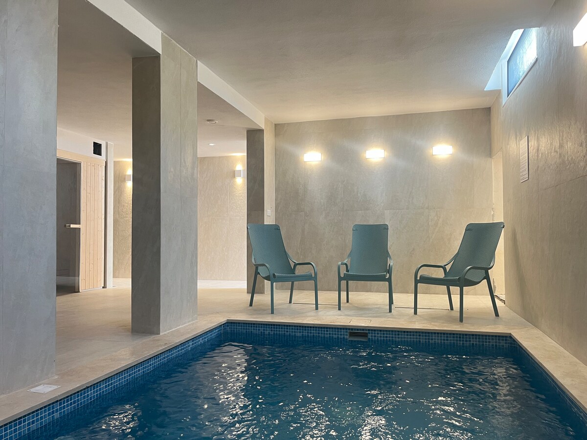 A serene indoor pool area features warm lighting and tiled walls. Three lounge chairs are positioned around the pool, providing a relaxing space for guests. Natural light filters through a window, adding to the calming atmosphere of the spa-like environment.