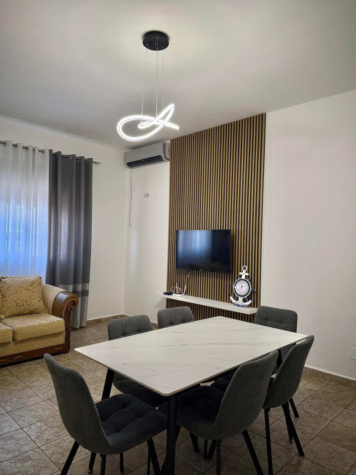 A modern dining area features a marble table surrounded by six gray chairs. A decorative light fixture hangs from the ceiling, illuminating the space. A cozy couch is positioned nearby, and a flat-screen television is mounted on an accent wall with wooden paneling.