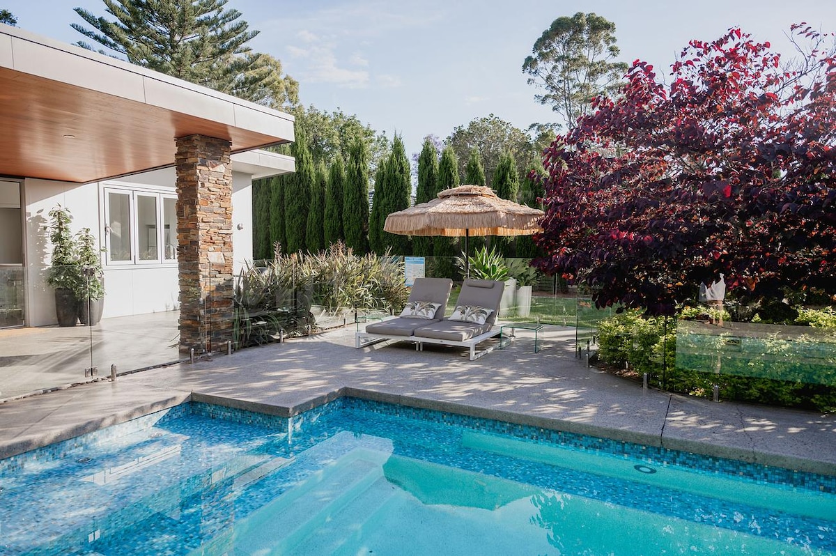 A serene outdoor space features a shimmering salt pool surrounded by landscaped greenery. Two lounge chairs are placed alongside the pool, complemented by a shaded cabana. Tall trees and colorful foliage create a tranquil atmosphere for relaxation.